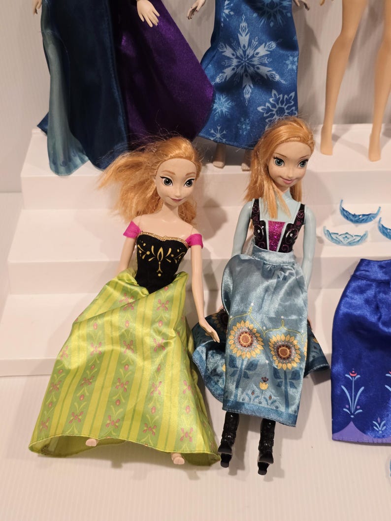 May include: Two Anna dolls from the film Frozen. One doll wears a black top with gold detailing and a green skirt with pink floral patterns. The other doll wears a blue dress with sunflower details. Other dolls and accessories are visible.