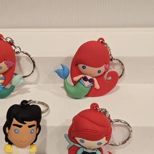 May include: Keychains featuring cartoon characters, including a mermaid with red hair and a blue tail, and a prince with black hair and a white and gold outfit. Each keychain has a silver metal ring.