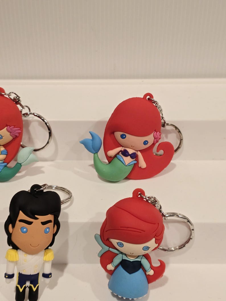 May include: Keychains featuring cartoon characters, including a mermaid with red hair and a blue tail, and a prince with black hair and a white and gold outfit. Each keychain has a silver metal ring.