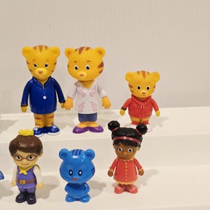 May include: A collection of six small plastic character figurines. The set includes two yellow tiger figures, one smaller yellow tiger, a figure with brown hair and glasses, a small blue cat-like figure, and a figure with dark hair in buns.