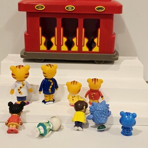 K&ouml;nnte beinhalten: Ein roter Spielzeug-Zugwagen mit gelbem Griff und mehreren kleinen Charakterfiguren. Die Figuren umfassen Tigercharaktere in verschiedenen Outfits und andere Charaktere in verschiedenen Farben. Der Spielzeug-Zugwagen hat drei Fenster.