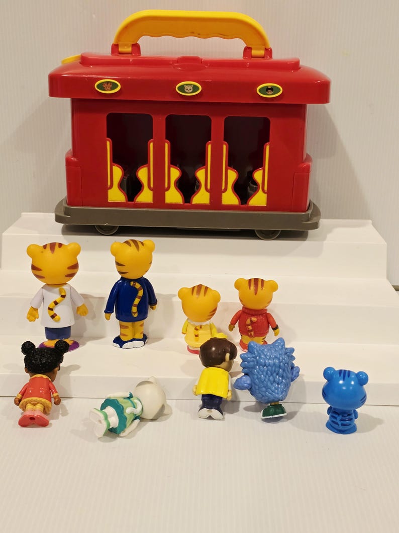 K&ouml;nnte beinhalten: Ein roter Spielzeug-Zugwagen mit gelbem Griff und mehreren kleinen Charakterfiguren. Die Figuren umfassen Tigercharaktere in verschiedenen Outfits und andere Charaktere in verschiedenen Farben. Der Spielzeug-Zugwagen hat drei Fenster.