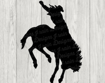 Könnte beinhalten: Schwarze Silhouette eines Cowboys, der einen sich aufbäumenden Bronco reitet. Die Silhouette ist vor einem Holzmaserungshintergrund platziert. Der Text "J RAIN DESIGNS" befindet sich am unteren Rand des Bildes.