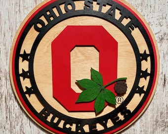 Może przedstawiać: Okrągły drewniany znak z czerwono-czarnym logo Ohio State Buckeyes. Logo przedstawia dużą czerwoną literę "O" z czarną obwódką i liściem buckeye w środku. Słowa "Ohio State" są napisane czarnym kolorem wokół górnej części znaku, a "Buckeyes" są napisane czarnym kolorem wokół dolnej części.