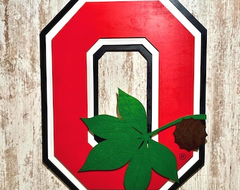 May include: A red and white wooden Ohio State University 'O' with a green buckeye leaf and a brown buckeye nut.