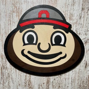 May include: A brown and tan cartoon face with a red and gray hat. The face has a big smile and large eyes. The hat has a white letter "O" on it.