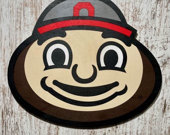 May include: A brown and tan cartoon face with a red and gray hat. The face has a big smile and large eyes. The hat has a white letter "O" on it.