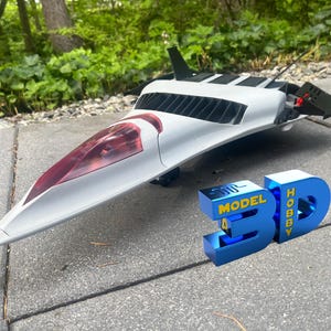 Pode incluir: Um modelo de vaivém espacial branco e preto com um cockpit tingido de vermelho. O modelo está sobre uma superfície cinzenta junto a um letreiro 3D azul que diz "3D MODEL HOBBY". O modelo tem asas pretas e um design detalhado.