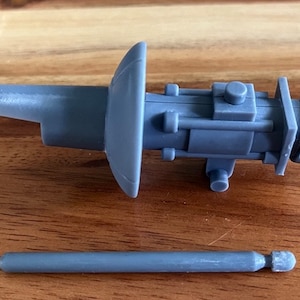 Puede incluir: Un modelo de motor de nave espacial de plástico gris con un cuerpo largo y cilíndrico y una boquilla redonda y plana. El motor tiene una pequeña varilla redonda de plástico gris unida a él.