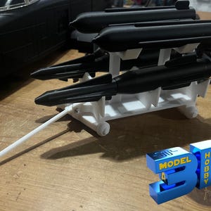 May include: A white plastic stand with four black missiles attached. The stand has a white handle and is on a wooden surface. The text "3D MODEL HOBBY" is visible in the lower right corner of the image.