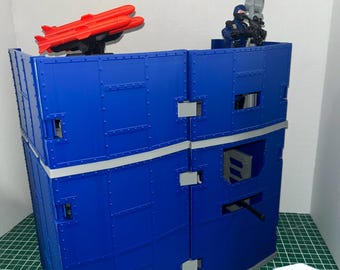 May include: A blue modular toy fortification wall with a gray trim, featuring a mounted red missile launcher on the left and a small blue action figure holding a gray weapon on the right. A 3D logo with the text MODEL 4 HOBBY is in the corner.