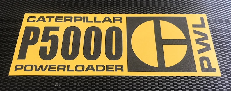 May include: A yellow and black sticker with the text "CATERPILLAR P5000 POWERLOADER". The Caterpillar logo is on the right, and the letters "PWL" are vertically aligned on the right edge. The sticker is rectangular in shape.