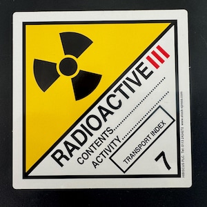 May include: A yellow and white warning sign with a black radiation symbol and the text "RADIOACTIVE III". The sign also includes the text "CONTENTS...", "ACTIVITY...", "TRANSPORT INDEX", and the number "7".