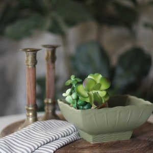May include: A green ceramic square bowl with a succulent plant inside. The bowl is sitting on a wooden cutting board with a striped cloth napkin next to it. Two brass candle holders are in the background.