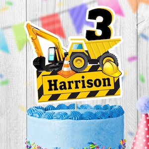 May include: A yellow construction vehicle cake topper with a black number 3, a construction worker's hat, and a sign that says "Harrison". The topper is on a blue frosted cake with colorful sprinkles.
