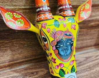 Pode incluir: Decoração de parede de cabeça de vaca de madeira pintada à mão com cores amarelo, laranja, rosa, verde e azul. O rosto da vaca apresenta um design detalhado com padrões florais e uma figura central com uma coroa.