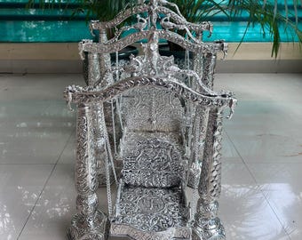 May include: Ornate silver-colored swing with intricate floral and geometric patterns. The swing features detailed carvings on the frame, seat, and backrest, suspended by chains. The decorative piece is likely for religious or ceremonial use.