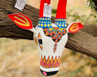 Pode incluir: Uma escultura de cabeça de vaca de madeira branca com detalhes pintados em vermelho, amarelo e azul. Os chifres são vermelhos com pontas amarelas e os olhos são pretos com pupilas brancas. A escultura está pendurada em um galho de árvore.