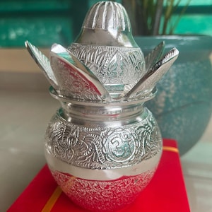 May include: A silver-colored, ornate container with a lotus-shaped lid. The container has detailed floral patterns and sits on a red surface. The design is likely for religious or decorative purposes.