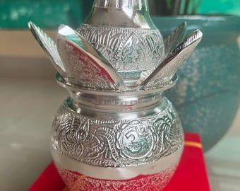 May include: A silver-colored, ornate container with a lotus-shaped lid. The container has detailed floral patterns and sits on a red surface. The design is likely for religious or decorative purposes.