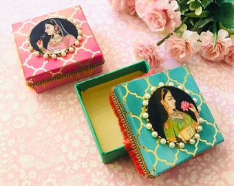 May include: Two square boxes with ornate designs. The boxes are decorated with images of women in traditional Indian clothing. The boxes are lined with a soft, fuzzy material. The boxes are a vibrant pink and teal.