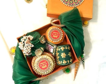 Op de afbeelding: Een groene en oranje geschenkdoos gevuld met verschillende items, waaronder een klein groen zakje, een ronde doos met de tekst "PRIYA KI MEHNDI", een groene en gouden armband en een klein groen en gouden decoratief item. De doos is versierd met een groene en gouden stof.