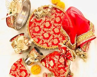 Könnte beinhalten: Ein traditionelles indisches Hochzeitszeremonien-Set mit rot-goldenen Mustern. Das Set enthält ein rundes Tablett, zwei kleine Schalen, eine Tasche, einen roten Schleier und dekorative Blumen.