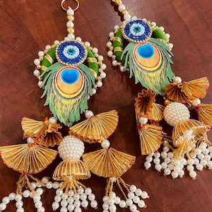 Könnte beinhalten: Zwei dekorative Türkranz-Anhänger mit einem Pfauenfeder-Design und einem blauen bösen Auge. Die Anhänger sind mit goldenen und weißen Perlen verziert und haben einen Quasten aus goldenem Stoff.