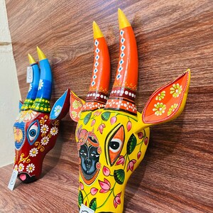 May include: Two wooden cow head sculptures with colourful painted details. The sculpture on the left is red, blue, and green with white flowers. The sculpture on the right is yellow with pink and green flowers and a blue face.
