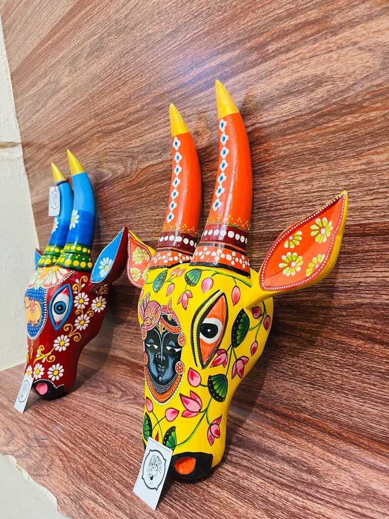 May include: Two wooden cow head sculptures with colourful painted details. The sculpture on the left is red, blue, and green with white flowers. The sculpture on the right is yellow with pink and green flowers and a blue face.