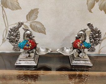 May include: Two silver peacock-shaped diyas with red, blue, and turquoise mosaic accents. The diyas are on a silver base and have a small bowl for oil and a wick.