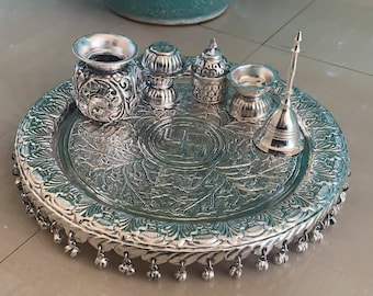 May include: A silver tray with a decorative border and a swastika symbol in the center. The tray is filled with small silver bowls and a silver bell.