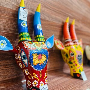 May include: Two colourful wooden cow head sculptures with intricate painted details. The sculpture on the left is red with blue horns and white flowers. The sculpture on the right is yellow with orange horns and white flowers.