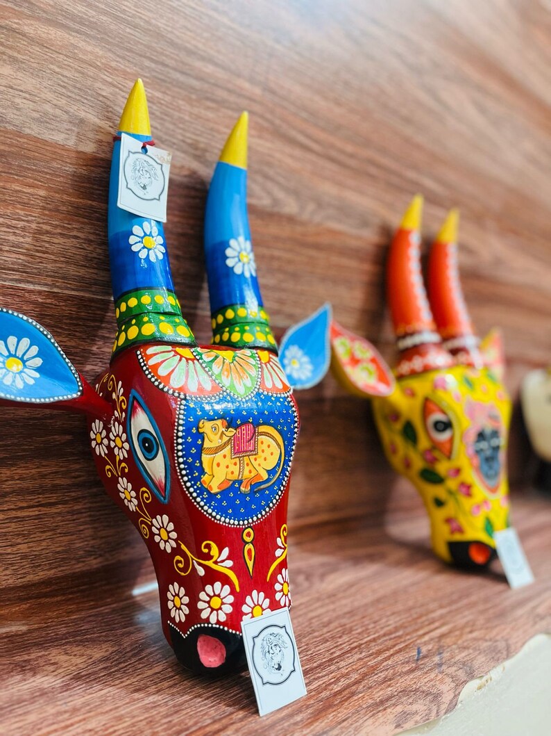 May include: Two colourful wooden cow head sculptures with intricate painted details. The sculpture on the left is red with blue horns and white flowers. The sculpture on the right is yellow with orange horns and white flowers.