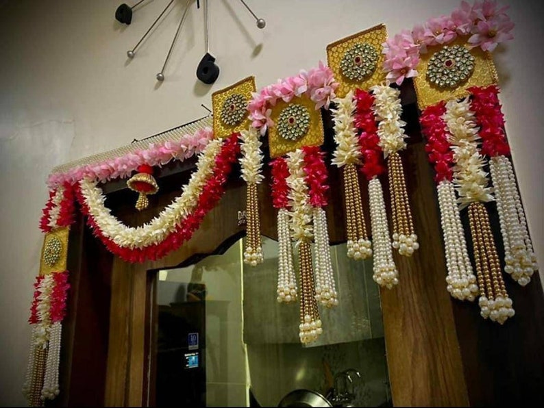May include: A traditional Indian door decoration with white and pink flower garlands, gold and pearl tassels, and gold-framed panels with jeweled accents.