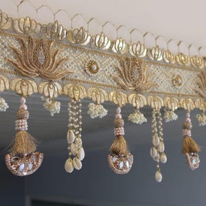May include: An ornate, cream-colored decorative valance with gold and pearl accents. The valance features lotus flower designs, beaded strands, tassels, and shell embellishments, creating an elegant, detailed aesthetic.
