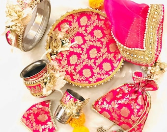 May include: A pink and gold Indian wedding ceremony set with a round tray, two small cups, a round metal container, a small pouch, and a pink veil. The set is decorated with gold floral patterns and pearls.
