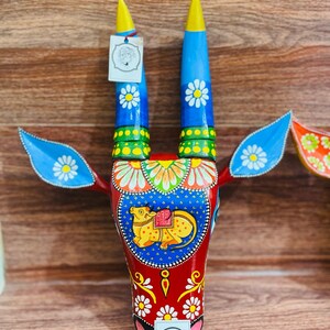 May include: A hand-painted wooden bull's head wall decoration featuring blue, red, yellow and green colours. The bull has white flowers painted on its horns and face. A small calf is painted on the bull's face.