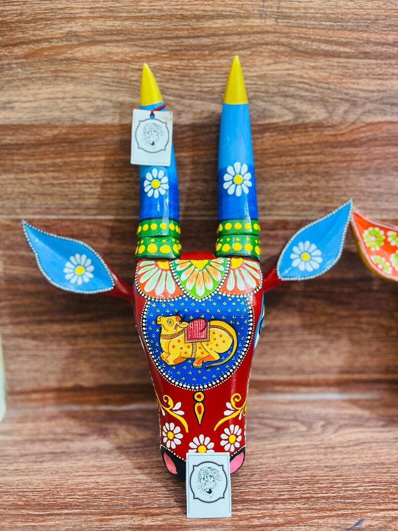 May include: A hand-painted wooden bull's head wall decoration featuring blue, red, yellow and green colours. The bull has white flowers painted on its horns and face. A small calf is painted on the bull's face.