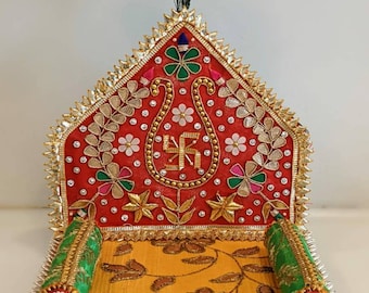 May include: A red, gold, and green decorated throne with a peacock feather on top. The throne has a yellow cushion with floral patterns and two green pillows. The throne is decorated with beads, sequins, and embroidery.