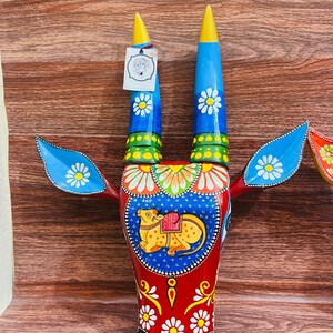 May include: A colourful wooden bull's head wall decoration featuring blue, red, yellow and green paint. The bull's horns are long and pointed, and the head is decorated with intricate floral patterns. The bull's face is painted with a yellow cow and a saddle.