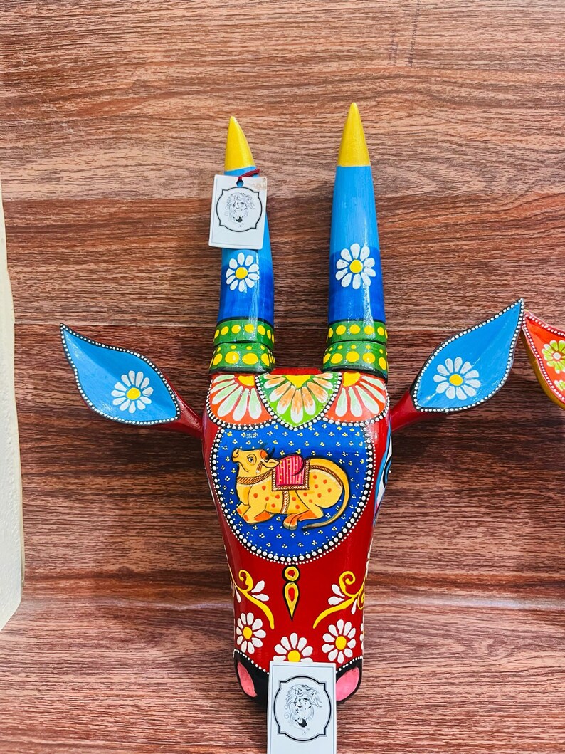 May include: A colourful wooden bull's head wall decoration featuring blue, red, yellow and green paint. The bull's horns are long and pointed, and the head is decorated with intricate floral patterns. The bull's face is painted with a yellow cow and a saddle.