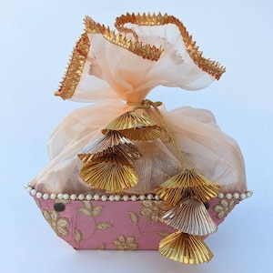 Può includere: Un cesto regalo rosa con una borsa in tessuto trasparente color pesca con nappine decorative dorate e argentate. Il cesto è decorato con un bordo di perle e ricami floreali.