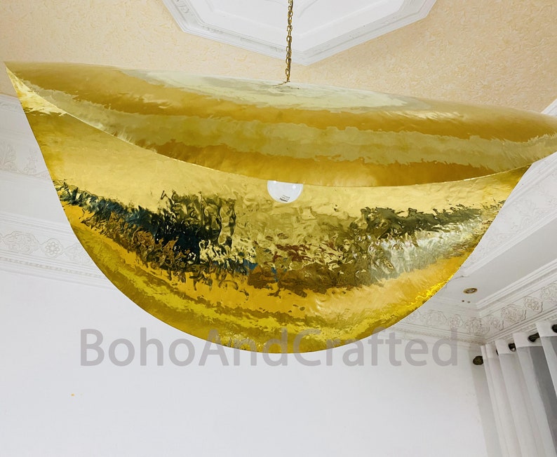 May include: A large, gold, hammered metal pendant light fixture hangs from the ceiling. The light fixture is shaped like a crescent moon and has a smooth, reflective surface.