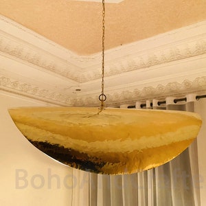 May include: A large, gold, hammered metal pendant light fixture hangs from a chain in a room with white walls and white curtains.