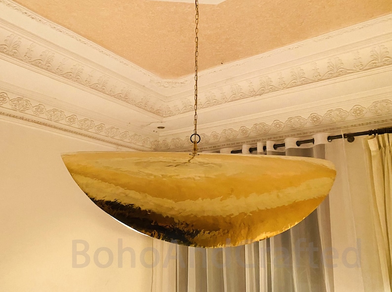 May include: A large, gold, hammered metal pendant light fixture hangs from a chain in a room with white walls and white curtains.