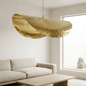 May include: A modern living room features a large, gold-colored, abstract pendant light fixture suspended from the ceiling. The light fixture is above a light-colored sofa and wooden coffee table. A large window reveals an outdoor zen garden.