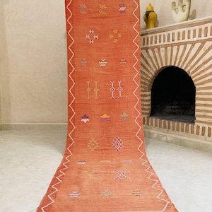 May include: A long, rust-colored Moroccan rug with intricate geometric patterns woven in shades of pink, purple, yellow, and white. The rug has a fringe border and is displayed against a white wall.