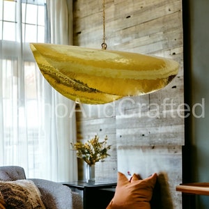 May include: A large, gold, hammered metal pendant light fixture hangs from the ceiling in a room with a wood wall and a sofa. The light fixture is shaped like a crescent moon.