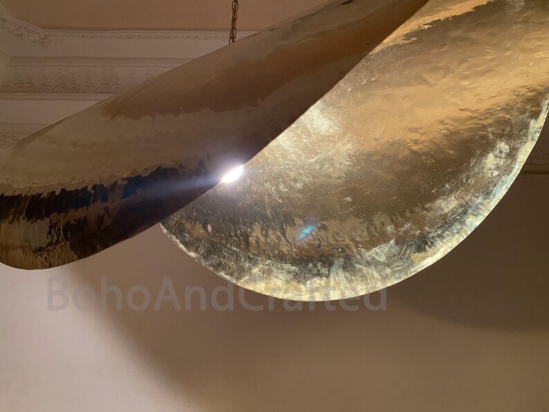 May include: A gold-toned metal pendant light fixture with two curved, overlapping panels. The light fixture is hanging from a chain.